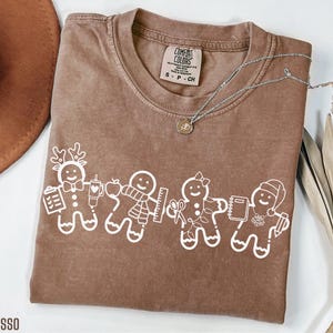 Könnte beinhalten: Espressofarbenes Comfort Colors T-Shirt mit weißer Lebkuchenmännchen-Grafik. Die Grafik zeigt ein Rentier, ein Lebkuchenmännchen mit einem Apfel, ein Lebkuchenmännchen mit einem Lineal und ein Lebkuchenmännchen mit einem Notizbuch. Eine silberne Halskette liegt auf dem Shirt.