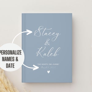 K&ouml;nnte beinhalten: Ein hellblaues Journal mit dem Text "Stacey & Kaleb" in wei&szlig;er Kursivschrift. Der Text "Two Hearts, One Journey" und das Datum "March 9, 2023" sind ebenfalls in wei&szlig;er Kursivschrift.