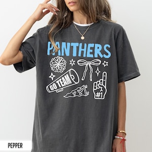 Puede incluir: Una camiseta de manga corta gris carb&oacute;n con el texto azul claro PANTHERS en la parte superior. Debajo del texto hay gr&aacute;ficos de l&iacute;neas blancas que incluyen un pomp&oacute;n, un lazo, un meg&aacute;fono con GO TEAM, un bander&iacute;n con corazones y un dedo de espuma #1.
