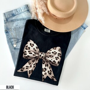 Puede incluir: Camiseta negra con un gran lazo de estampado de leopardo. La camiseta est&aacute; puesta en una modelo que lleva pantalones vaqueros azules y un sombrero marr&oacute;n.