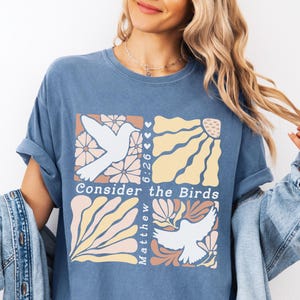 Op de afbeelding: Een blauw T-shirt met een grafisch ontwerp van twee witte duiven met bloemen- en zonmotieven. De tekst "Consider the Birds" en "Matthew 6:26" zijn opgenomen in het ontwerp.