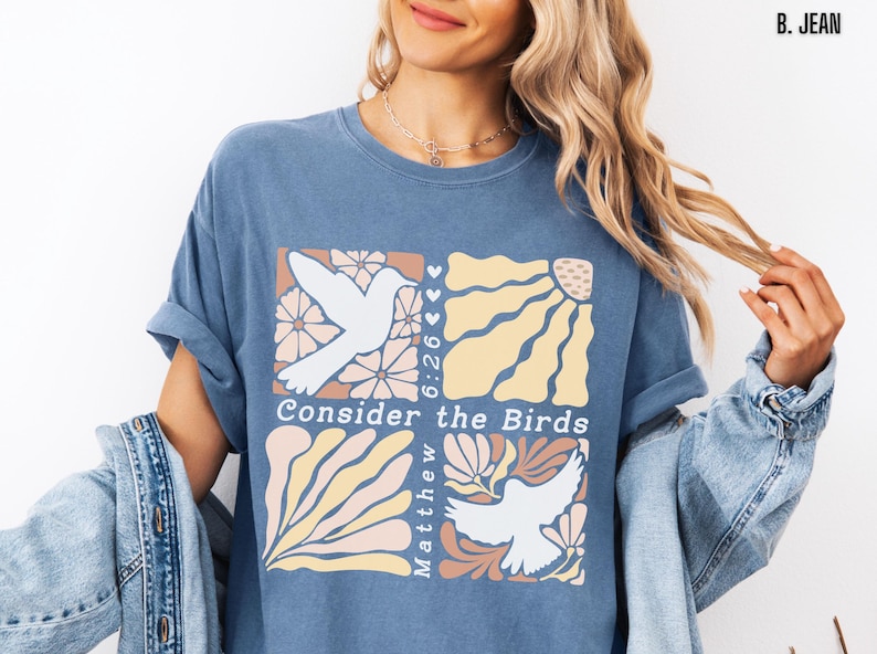 Op de afbeelding: Een blauw T-shirt met een grafisch ontwerp van twee witte duiven met bloemen- en zonmotieven. De tekst "Consider the Birds" en "Matthew 6:26" zijn opgenomen in het ontwerp.