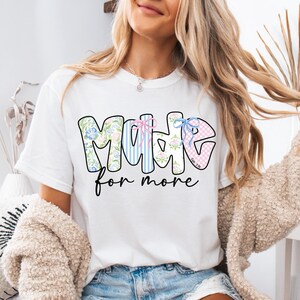 Op de afbeelding: Wit T-shirt met de tekst "MOMME" in een bloemen-, gestreept en ruitpatroon, met de woorden "for more" in een scriptlettertype. Het shirt heeft een ronde hals en korte mouwen.