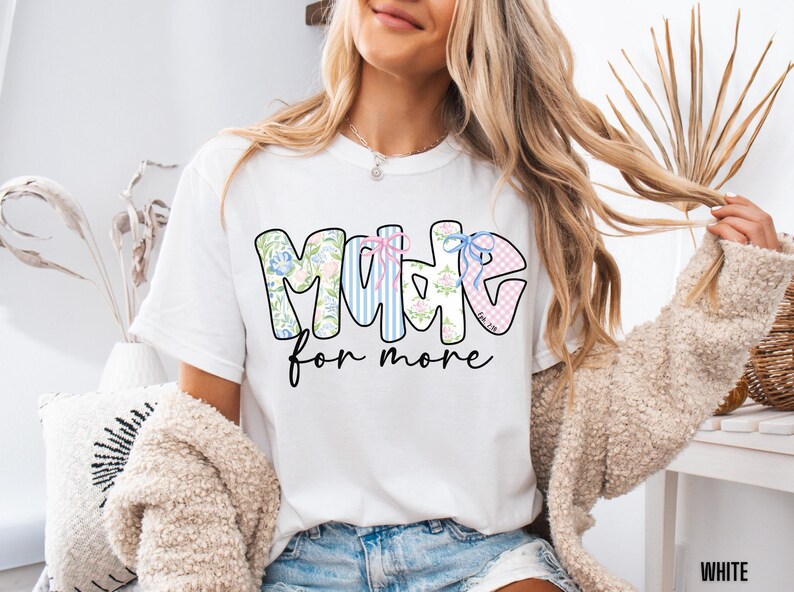 Op de afbeelding: Wit T-shirt met de tekst "MOMME" in een bloemen-, gestreept en ruitpatroon, met de woorden "for more" in een scriptlettertype. Het shirt heeft een ronde hals en korte mouwen.