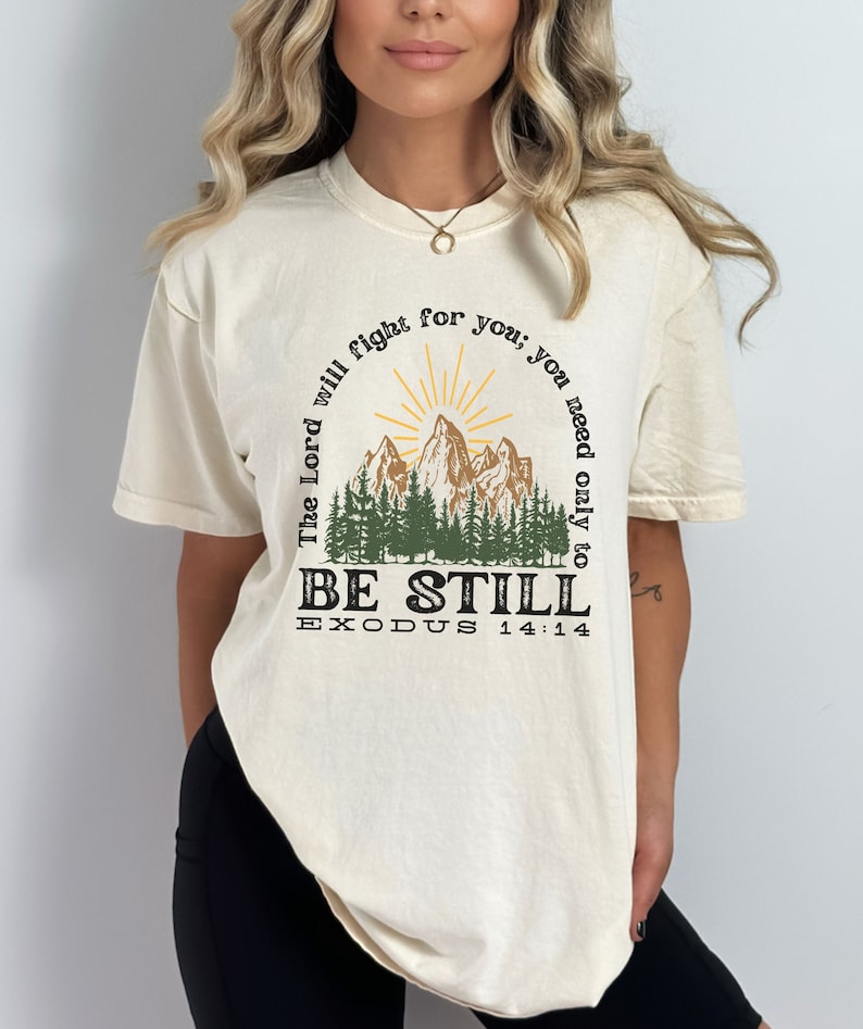 May include: A white t-shirt with a graphic design of a mountain range with a sunrise behind it. The text "The Lord will fight for you; you need only to be still" is written around the mountain range. The text "Exodus 14:14" is written below the mountain range.