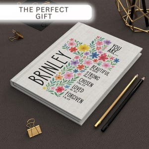 May include: A white journal with the name "BRINLEY" and the words "YOU ARE... BEAUTIFUL, STRONG, CHOSEN, LOVED, FORGIVEN" printed on the cover. The cover features a floral cross design with colorful flowers. Two pencils and a gold paper clip are also visible.