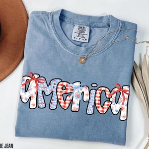 May include: A blue jean t-shirt with the word "America" written in white outlined letters. The letters are decorated with red and white gingham, red and white fireworks, and red bows.
