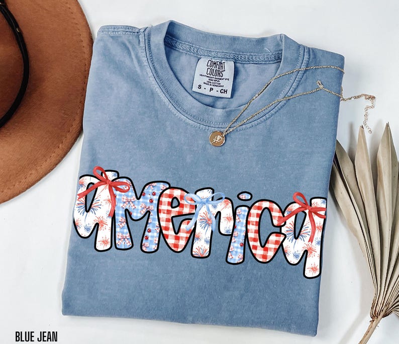 May include: A blue jean t-shirt with the word "America" written in white outlined letters. The letters are decorated with red and white gingham, red and white fireworks, and red bows.