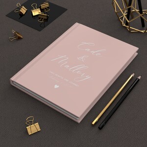 K&ouml;nnte beinhalten: Ein rosafarbenes Hardcover-Journal mit dem Text "Cade & Mallory" in wei&szlig;er Kursivschrift. Der Text "Two Hearts, One Journey" ist darunter in wei&szlig; mit einem kleinen wei&szlig;en Herzsymbol gedruckt.