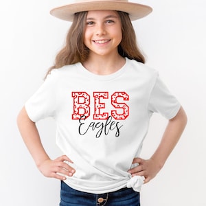 May include: White t-shirt with red and white "BES Eagles" text on the front. The shirt is being worn by a young person wearing a brown hat.