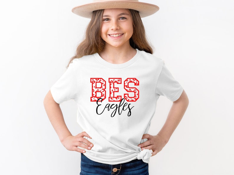 May include: White t-shirt with red and white "BES Eagles" text on the front. The shirt is being worn by a young person wearing a brown hat.