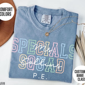 May include: A blue jean coloured t-shirt with the words "SPECIALS SQUAD Ms. Collins P.E." in colourful block letters. The shirt has a crew neck and short sleeves. A gold necklace with a pendant is draped across the shirt.