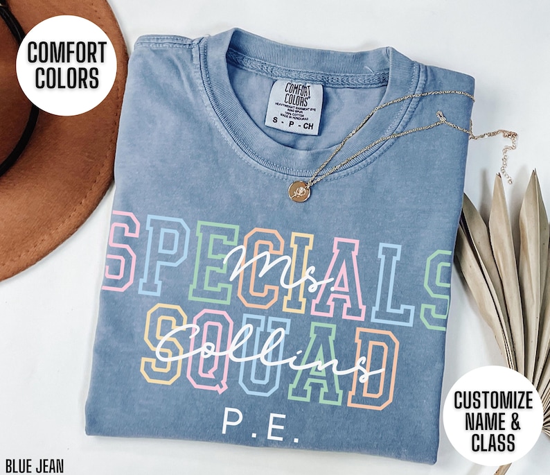 May include: A blue jean coloured t-shirt with the words "SPECIALS SQUAD Ms. Collins P.E." in colourful block letters. The shirt has a crew neck and short sleeves. A gold necklace with a pendant is draped across the shirt.