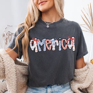 May include: A dark gray t-shirt with a red, white, and blue "America" graphic. The graphic is made up of stars, stripes, and gingham patterns.