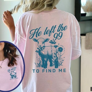 May include: A pale pink t-shirt featuring the teal text "He left the 99 TO FIND ME" and a lamb illustration with flowers. The text "Luke 15:4-7" is also included. The shirt is a relaxed fit, ideal for everyday wear.