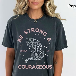 May include: A dark gray t-shirt with a white tiger graphic and the text "Be Strong & Courageous" in pink lettering. The text "Joshua 1:9" is also included in white lettering.