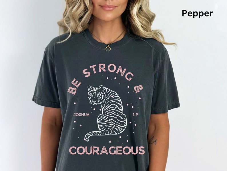 May include: A dark gray t-shirt with a white tiger graphic and the text "Be Strong & Courageous" in pink lettering. The text "Joshua 1:9" is also included in white lettering.