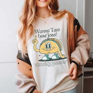 May include: A white sweatshirt with a graphic of a taco with arms and legs. The taco is pointing up with its index finger and has the text "Wanna Taco bout Jesus?" printed on the shirt. The text "FOR GOD SO LOVED THE WORLD THAT HE GAVE HIS ONE AND ONLY SON, THAT WHOEVER BELIEVES IN HIM SHALL NOT PERISH BUT HAVE ETERNAL LIFE JOHN 3:16" is printed below the taco graphic.