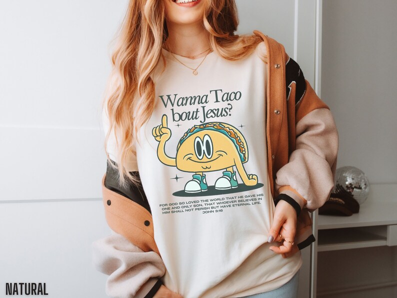 May include: A white sweatshirt with a graphic of a taco with arms and legs. The taco is pointing up with its index finger and has the text "Wanna Taco bout Jesus?" printed on the shirt. The text "FOR GOD SO LOVED THE WORLD THAT HE GAVE HIS ONE AND ONLY SON, THAT WHOEVER BELIEVES IN HIM SHALL NOT PERISH BUT HAVE ETERNAL LIFE JOHN 3:16" is printed below the taco graphic.