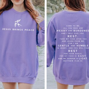May include: Purple sweatshirt with a white bird graphic on the front and a white text quote from the Bible on the back. The quote reads "Come to me, all you who are weary and burdened, and I will give you rest. Take my yoke upon you and learn from me, for I am gentle and humble in heart, and you will find rest for your souls. For my yoke is easy and my burden is light. Matthew 11:28-30".