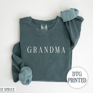 May include: A teal blue sweatshirt with the word "GRANDMA" printed in white on the front. The sweatshirt is folded in half and the sleeves are rolled up. The text "DTG PRINTED" is visible in the lower right corner of the image.