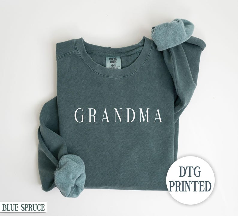 May include: A teal blue sweatshirt with the word "GRANDMA" printed in white on the front. The sweatshirt is folded in half and the sleeves are rolled up. The text "DTG PRINTED" is visible in the lower right corner of the image.