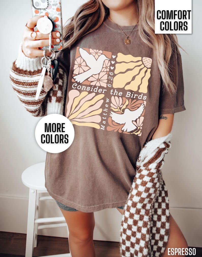 Op de afbeelding: Een bruin T-shirt met een grafisch ontwerp met twee witte duiven, bloemen en de tekst "Consider the Birds Matthew 6:26".