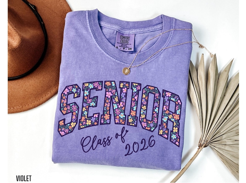 Comfort Colors shirt featuring the word 'Senior' in large, colorful floral print with 'Class of 2026' written underneath in a clean, modern font. Available in multiple styles including t-shirt, long sleeve, tank top, sweatshirt, and youth sizes. Displayed in soft, trendy colors like Ivory, Blossom, Violet, Chalky Mint, and Butter. Perfect for senior year celebrations, school events, senior photos, and graduation gifts.