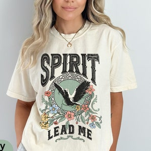 K&ouml;nnte beinhalten: Ein cremefarbenes T-Shirt mit einem Vintage-Grafikdesign, das einen schwarzen Vogel mit floralen Akzenten zeigt. Der Text "SPIRIT LEAD ME" ist auf das Shirt gedruckt.