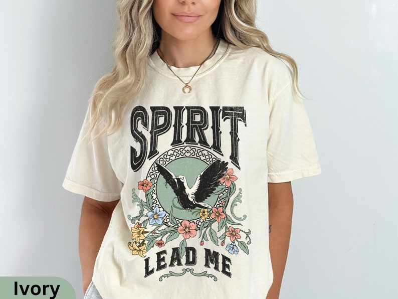 K&ouml;nnte beinhalten: Ein cremefarbenes T-Shirt mit einem Vintage-Grafikdesign, das einen schwarzen Vogel mit floralen Akzenten zeigt. Der Text "SPIRIT LEAD ME" ist auf das Shirt gedruckt.