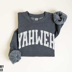 May include: Dark grey heather sweatshirt with the word "YAHWEH" printed in white, arched across the chest. The sleeves are rolled up. The words "DARK GREY HEATHER" are printed in black at the bottom left.
