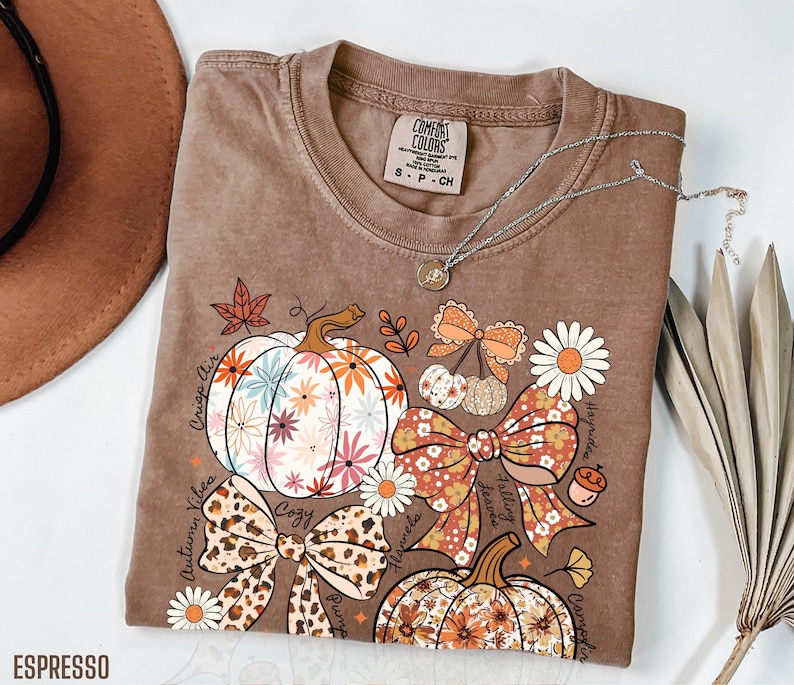 Comfort Colors t-shirt, long sleeve, lightweight sweatshirt, or youth tee with a coquette fall design featuring floral bows, floral pumpkins, autumn leaves, and acorns. The design is in soft, warm fall colors printed on fall-toned shirts like Moss, Yam, Terracotta, Ivory, Espresso, and Pepper. A cozy and cute seasonal shirt perfect for autumn lovers, fall photos, pumpkin patch visits, and Thanksgiving gatherings. Available in adult and youth sizing for a fun fall outfit.