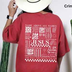 May include: A red t-shirt with a white graphic design featuring the word "JESUS" surrounded by other words and symbols. The text includes "Name Above All Names", "Son of God", "Holy One", "Redeemer", "Counselor", "Deliverer", "I Am", "Savior", "The Vine", "Prince of Peace", "Everlasting Father", "The Way The Truth The Life".