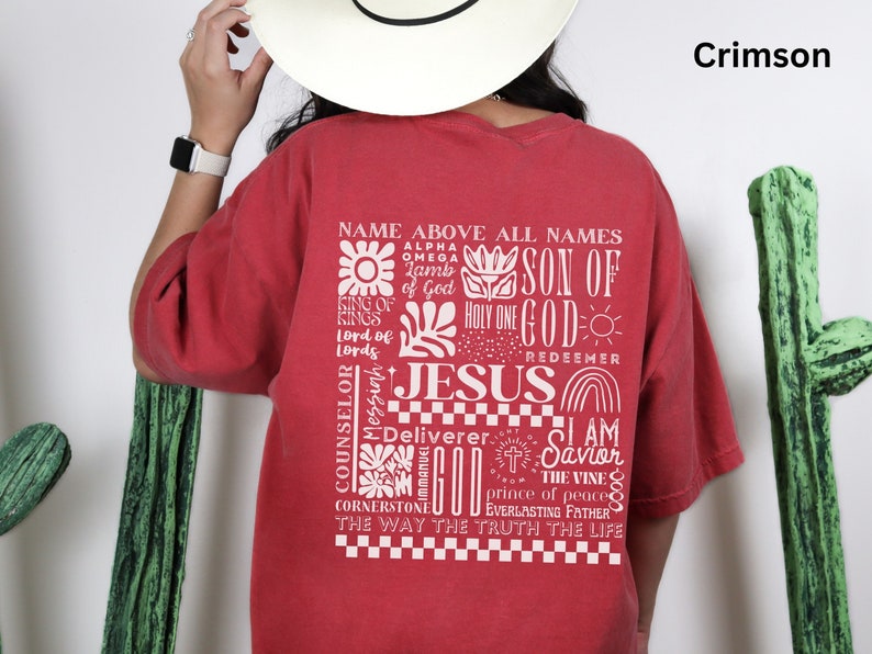 May include: A red t-shirt with a white graphic design featuring the word "JESUS" surrounded by other words and symbols. The text includes "Name Above All Names", "Son of God", "Holy One", "Redeemer", "Counselor", "Deliverer", "I Am", "Savior", "The Vine", "Prince of Peace", "Everlasting Father", "The Way The Truth The Life".