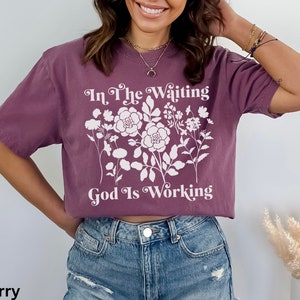 Op de afbeelding: Een paars gecropped t-shirt met een wit bloemenmotief en de tekst "In The Waiting God Is Working".