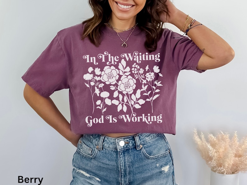 Op de afbeelding: Een paars gecropped t-shirt met een wit bloemenmotief en de tekst "In The Waiting God Is Working".