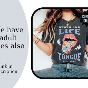 Pu&ograve; includere: T-shirt grigio scuro con le parole "DEATH AND LIFE" e "TONGUE" in stile vintage. La grafica della lingua presenta fiori e fulmini. Il testo "We have adult sizes also" &egrave; a sinistra.