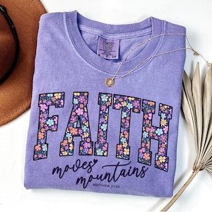 May include: Violet Comfort Colors t-shirt with the word "FAITH" in a floral design. The text "moves mountains" and Matthew 17:20 are below the word "FAITH". A gold necklace and a brown hat are next to the shirt.