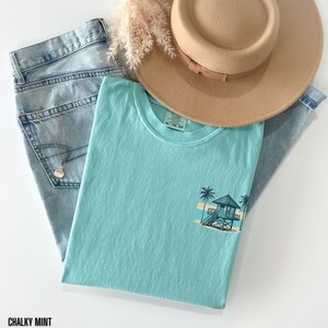 Puede incluir: Una camiseta azul claro con un gr&aacute;fico de una escena de playa con una caseta de salvavidas y palmeras. El texto "Chalky Mint" es visible en la parte inferior de la imagen.