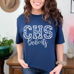 May include: Navy blue t-shirt with the white text "CHS Bobcats". The letters "CHS" are filled with a leopard print pattern. The shirt is a crew neck style.