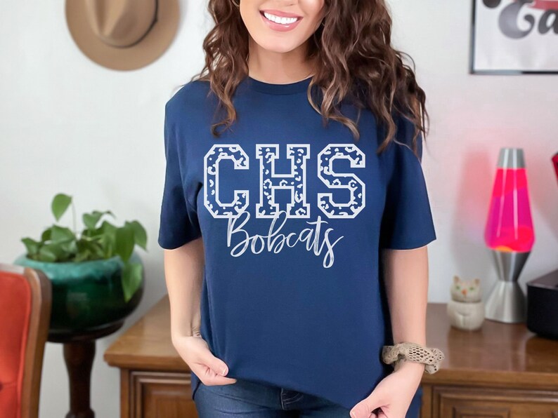 May include: Navy blue t-shirt with the white text "CHS Bobcats". The letters "CHS" are filled with a leopard print pattern. The shirt is a crew neck style.