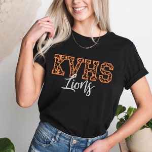 May include: Black t-shirt with a white and orange "KVHS Lions" graphic. The letters are outlined in black and filled with a leopard print pattern.