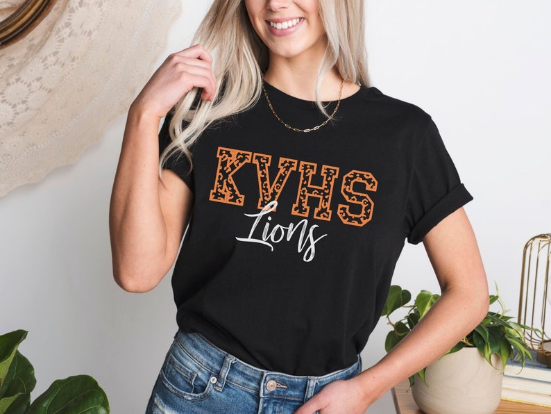 May include: Black t-shirt with a white and orange "KVHS Lions" graphic. The letters are outlined in black and filled with a leopard print pattern.