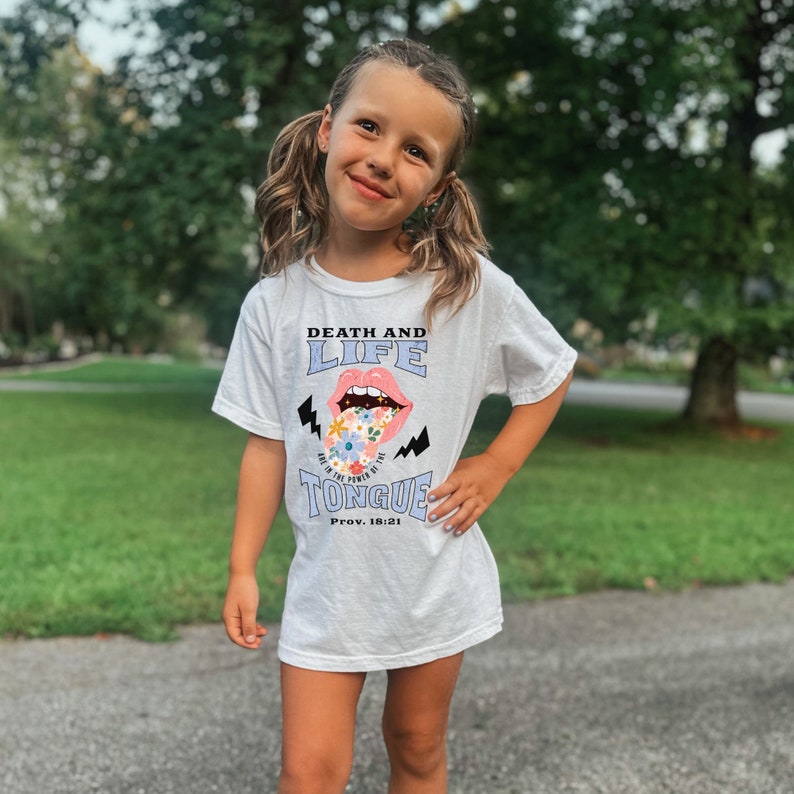 Pu&ograve; includere: T-shirt bianca con la scritta "DEATH AND LIFE ARE IN THE POWER OF THE TONGUE" e una grafica di una bocca con fiori. La maglietta presenta anche il versetto biblico "Prov. 18:21".