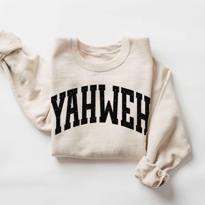 May include: A sand-colored sweatshirt with the word "YAHWEH" printed in black, arched lettering. The sweatshirt is folded, with the sleeves rolled up. The word "SAND" is printed in black at the bottom left corner.