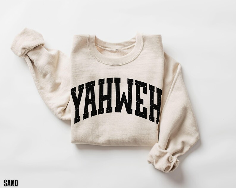 May include: A sand-colored sweatshirt with the word "YAHWEH" printed in black, arched lettering. The sweatshirt is folded, with the sleeves rolled up. The word "SAND" is printed in black at the bottom left corner.