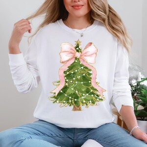 K&ouml;nnte beinhalten: Wei&szlig;es Langarm-Sweatshirt mit einem Aquarell-Weihnachtsbaum-Design. Der Baum ist gr&uuml;n mit goldenen Akzenten, einer rosa Schleife und Lichterketten. Das Wort "WHITE" ist unten aufgedruckt.