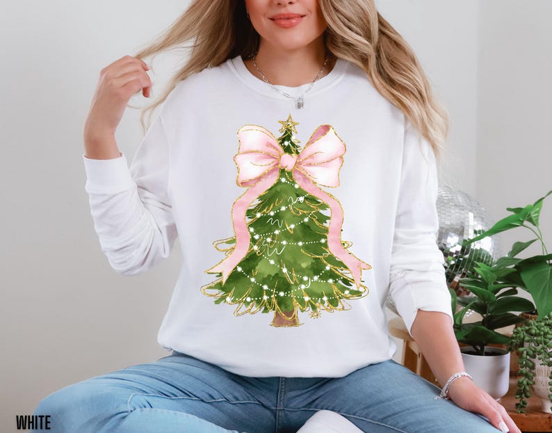 K&ouml;nnte beinhalten: Wei&szlig;es Langarm-Sweatshirt mit einem Aquarell-Weihnachtsbaum-Design. Der Baum ist gr&uuml;n mit goldenen Akzenten, einer rosa Schleife und Lichterketten. Das Wort "WHITE" ist unten aufgedruckt.