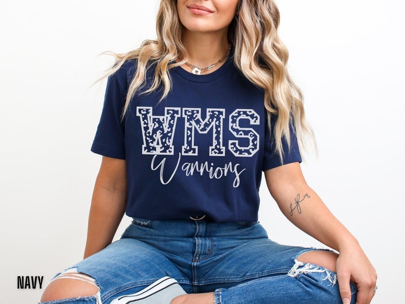 May include: Navy blue t-shirt with the text "WMS Warriors" in white. The letters "WMS" are filled with a leopard print design. The shirt is a short-sleeved crew neck.