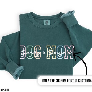 May include: A teal blue sweatshirt with the text "DOG MOM" in colorful block letters. The word "Dog" is in pink, yellow, and green. The word "Mom" is in blue, light blue, and pink. The text "Darby & Regan" is written in cursive in white. The text "ONLY THE CURSIVE FONT IS CUSTOMIZABLE" is written in white at the bottom of the image.